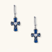 Silver huggie earrings with blue enamel cross charms featuring bat wings and heart-shaped crystal center