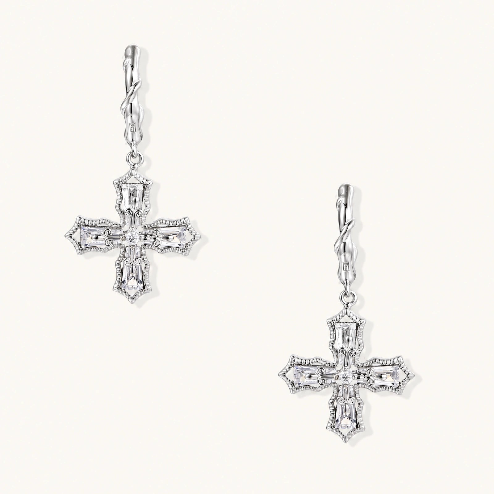 Ornate white gold-plated cross earrings with baguette-cut zirconia stones and twisted leverback hooks on beige background