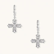 Ornate white gold-plated cross earrings with baguette-cut zirconia stones and twisted leverback hooks on beige background