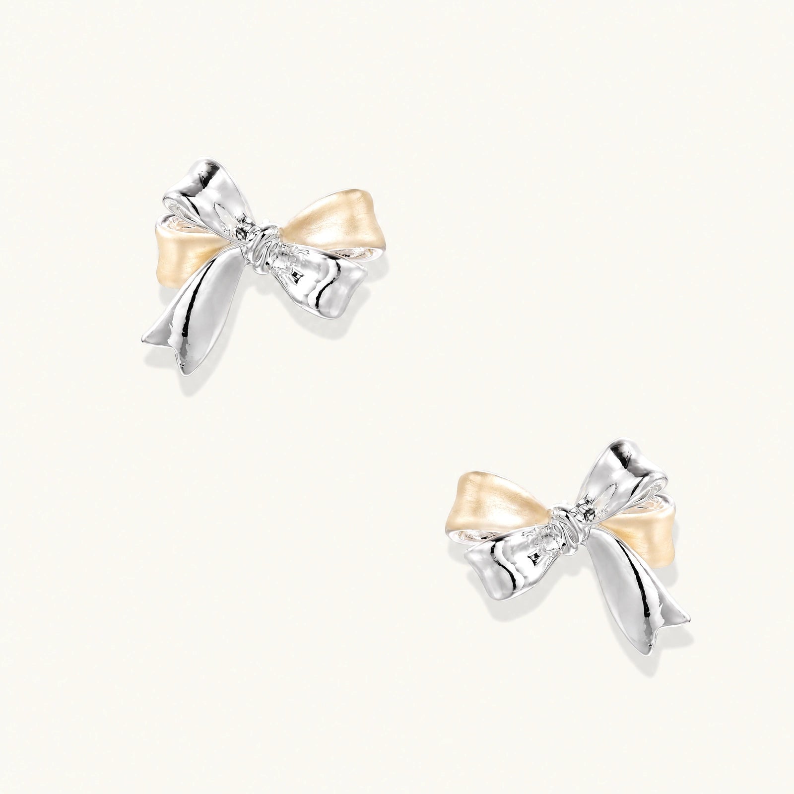 Pair of bow stud earrings with mirror-finish silver-tone ribbons and cream enamel-look loops on a cream background.