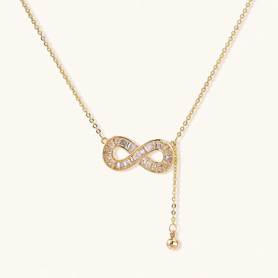 Gold infinity pendant with baguette crystals on adjustable chain against cream background.