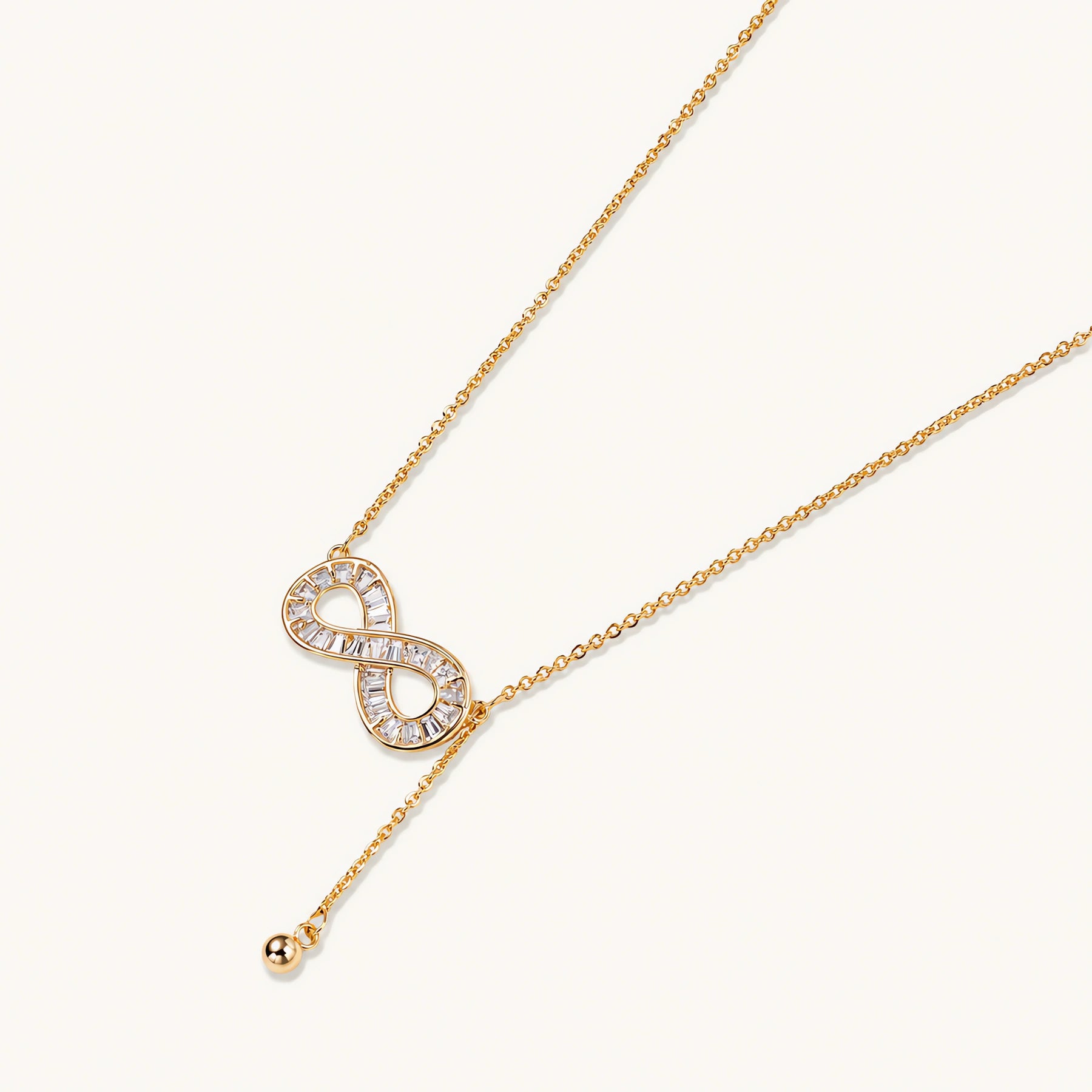 Gold infinity pendant with baguette crystals on adjustable chain against cream background.