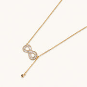 Gold infinity pendant with baguette crystals on adjustable chain against cream background.