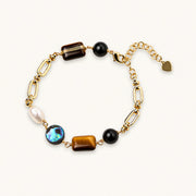 Gold chain bracelet with tiger's eye, obsidian, pearl, and iridescent abalone stones on cream background.