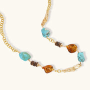 Gold plated eco-brass chain necklace with turquoise nuggets, amber-toned stones, brown discs and molten gold accents on cream.