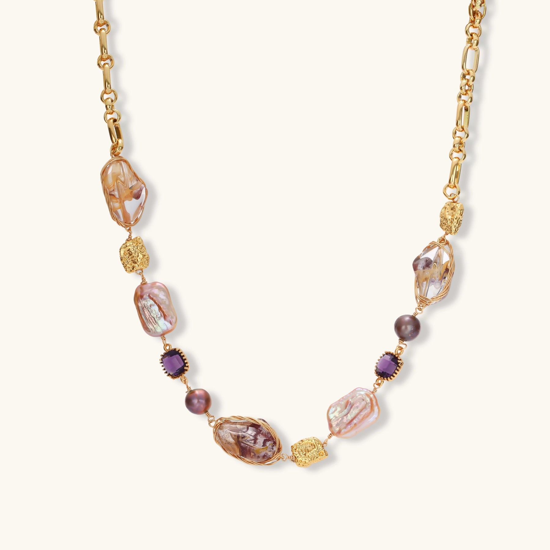 Gold plated chain necklace with wire-wrapped baroque pearls, purple faceted stones and textured gold beads on a cream background.