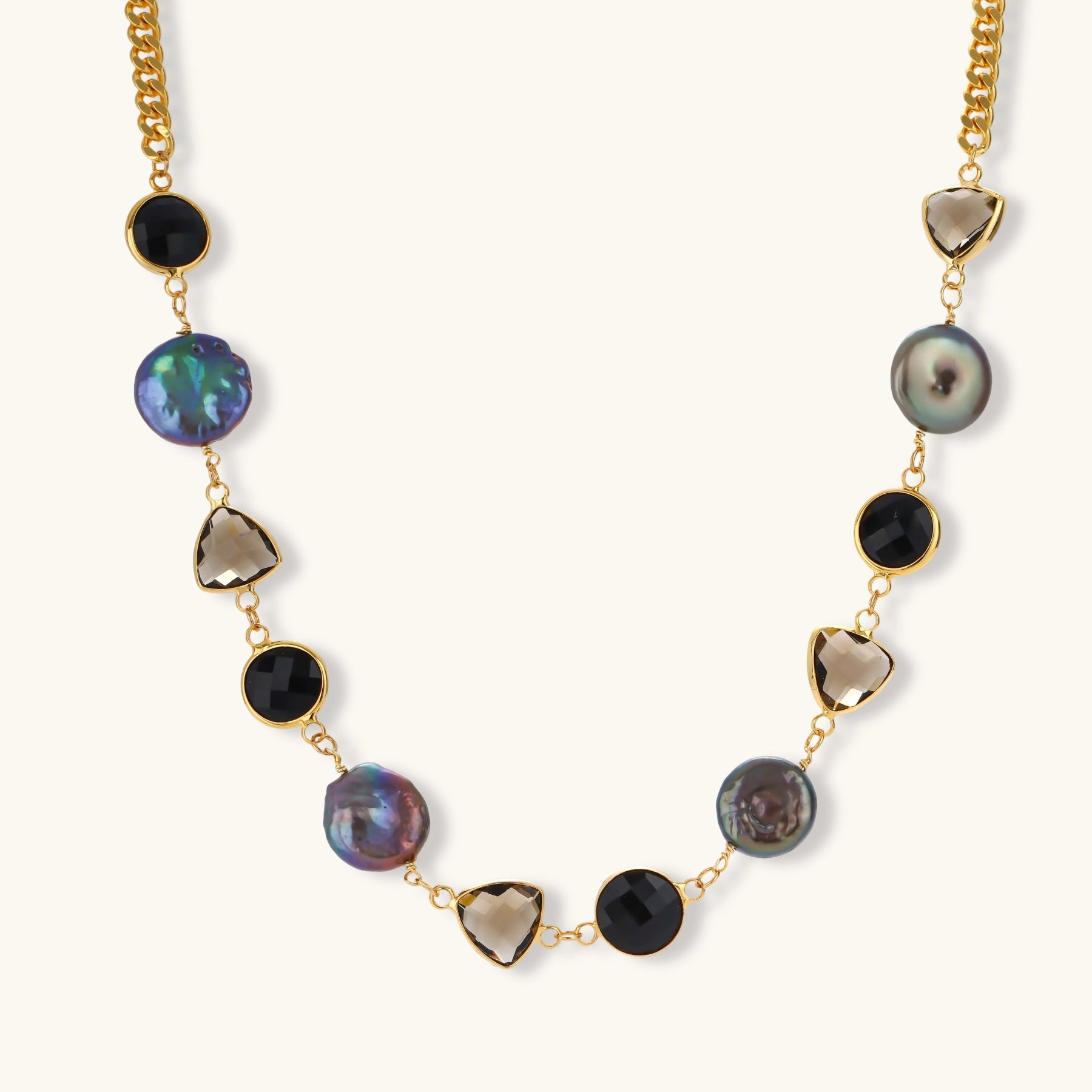 Gold plated curb chain necklace with iridescent baroque pearls, black round stones and smoky triangular stones on a cream background.