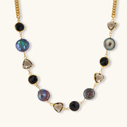 Gold plated curb chain necklace with iridescent baroque pearls, black round stones and smoky triangular stones on a cream background.