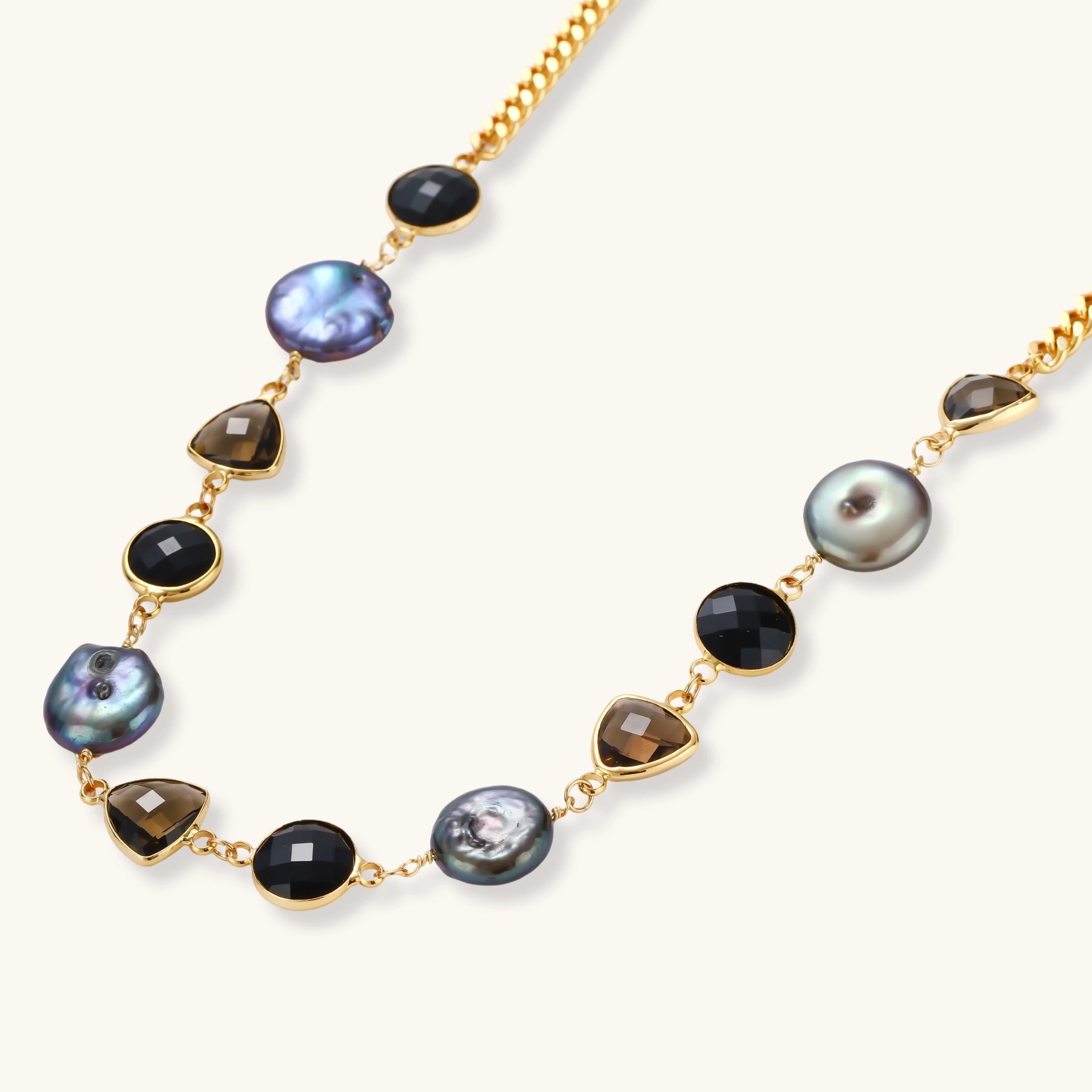 Gold plated curb chain necklace with iridescent baroque pearls, black round stones and smoky triangular stones on a cream background.