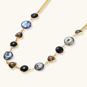 Gold plated curb chain necklace with iridescent baroque pearls, black round stones and smoky triangular stones on a cream background.