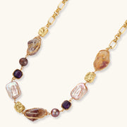 Gold plated chain necklace with wire-wrapped baroque pearls, purple faceted stones and textured gold beads on a cream background.